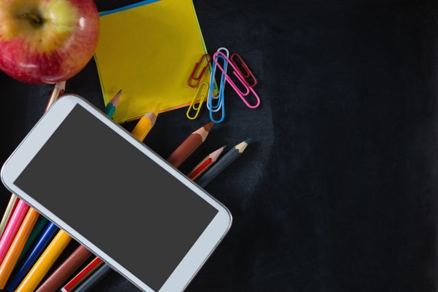School supplies including pencils, paper clips, sticky notes, and an apple are arranged next to a smartphone on a black background. This image is ideal for back-to-school promotions, educational content, and technology in education themes.