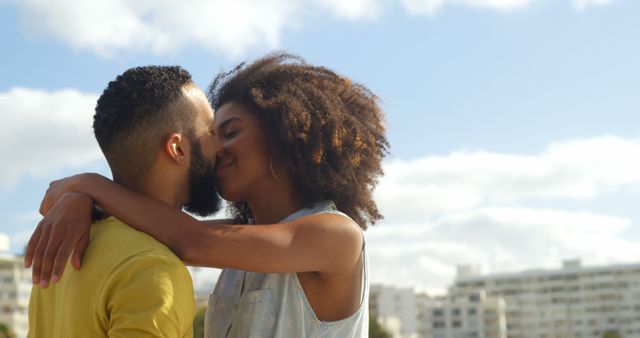 This close-up shot features a romantic couple embracing and about to kiss with a modern cityscape in the background and a clear, blue sky. Perfect for use in relationship advice articles, love-themed social media posts, or advertisements promoting romantic getaways and dating services.