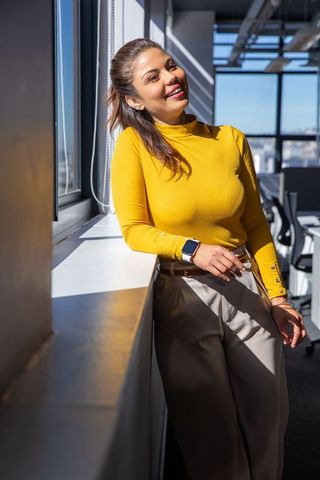 Smiling professional woman leaning on sunlit office windowsill in mustard top with smartwatch
