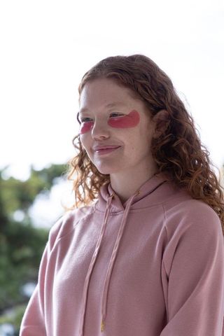 Young woman wearing under-eye gel patches and pink hoodie standing outdoors smiling