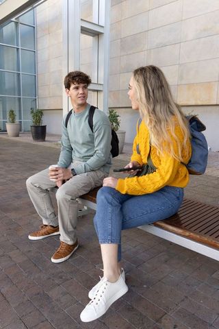 College students chatting on campus bench holding coffee and smartphone, casual backpacks
