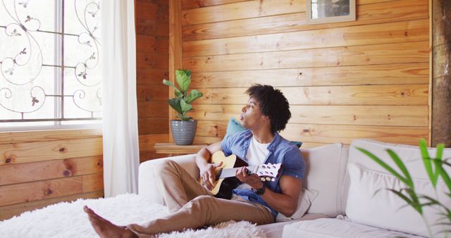 This picture depicts a young man relaxing on a cozy couch while playing an acoustic guitar in a rustic living room filled with natural light. The wooden interior creates a warm and inviting atmosphere. Ideal for use in articles and advertisements related to lifestyle, music, relaxation, homemaking, or creative hobbies.