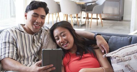Diverse couple lounging on couch sharing tablet together in bright modern cozy living room