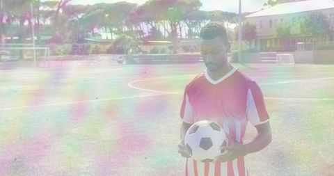 Mid-adult male holding soccer ball on sunlit football pitch wearing red-striped jersey