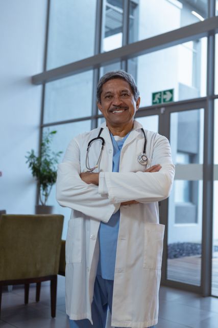 Mature male doctor standing confidently in hospital lobby with arms crossed, smiling. Ideal for healthcare, medical services, hospital promotions, and professional healthcare staff imagery.