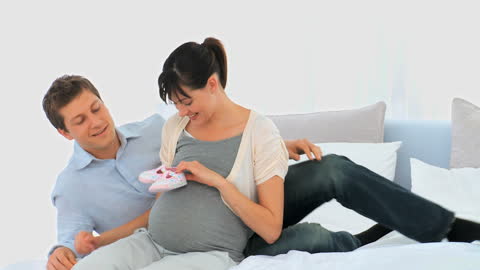 Expecting Parents Smiling and Holding Baby Shoes in Bedroom