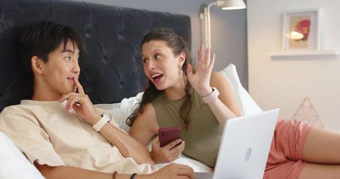 Mixed-race couple relaxing on bed, chatting and waving during video call with devices
