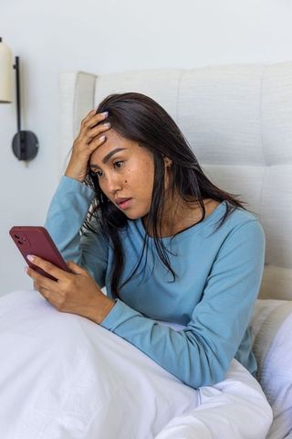 Asian woman checking smartphone and looking worried in bed wearing teal loungewear