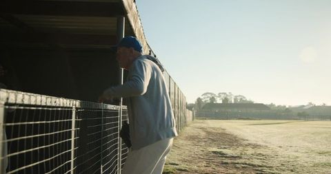 Senior man leaning on fence watching frosty sports field at dawn with blue cap and bag