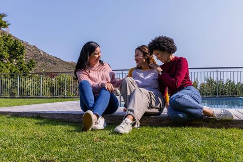 Girlfriends laughing and chatting on raised wooden pool deck in sunny backyard