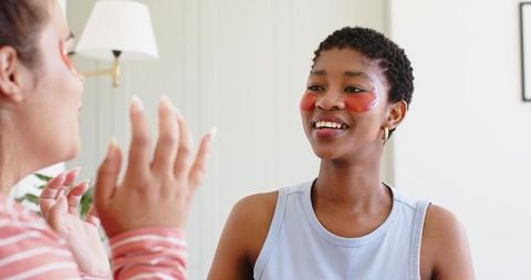Smiling woman wearing red hydrogel eye patches enjoying skincare moment with friend