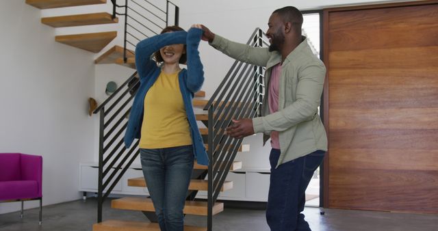 A cheerful couple dancing together in a stylish living room with a modern staircase. The man playfully spins the woman as they enjoy a fun moment. Perfect for depicting relationships, joy, and moments shared at home. Ideal for advertisements, family-oriented content, and home lifestyle materials.