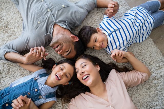 A family of four is lying on a carpeted floor, holding hands and smiling at each other. Both parents and children appear joyful and content, enjoying a moment of togetherness. Ideal for use in advertisements or articles about family bonding, home life, happiness, or parenting.