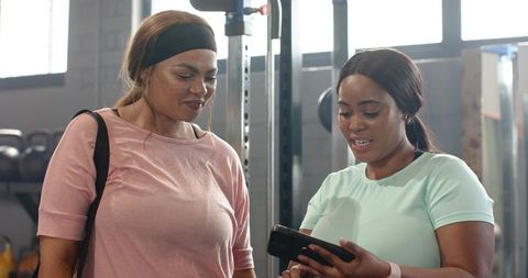 Diverse women comparing workout plan on smartphone in sunlit gym weight area
