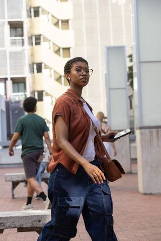 African-american woman walking city plaza holding phone and tablet casual urban style