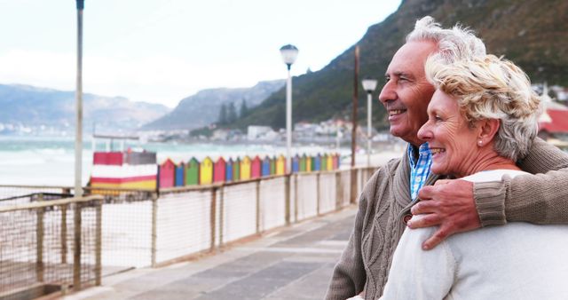A senior Caucasian couple enjoys a scenic view at a beachfront, with colorful beach huts in the background and copy space. Their warm embrace and smiles suggest a moment of contentment and companionship.