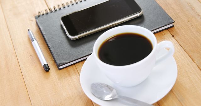 A cup of coffee sits next to a smartphone and a notebook on a wooden table, with copy space. Ideal for themes of morning routines, work breaks, or planning sessions.