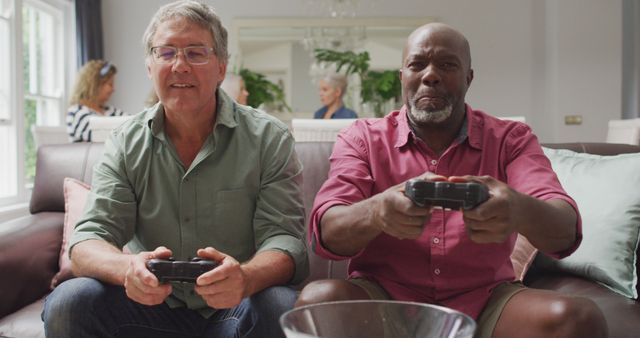 Senior men sitting on couch in living room, holding game controllers, enjoying video game. Friends in background having conversation. Perfect for illustrating active aging, friendship, and leisure activities for seniors.