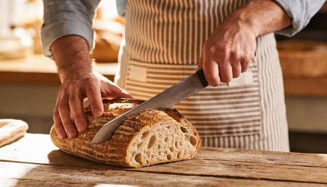 Baker slicing rustic artisan sourdough loaf on wooden countertop wearing striped apron
