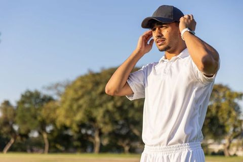 Indian cricketer adjusting cap on sunlit grass field wearing white kit and smartwatch
