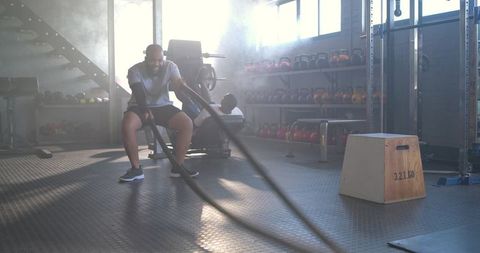 African american man training with battle ropes in backlit industrial gym for strength