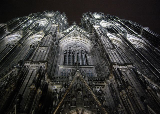 An upward view of a Gothic cathedral's intricately detailed facade illuminated at night against a dark sky. This image can be used for articles, blogs or websites focusing on historical architecture, travel destinations, cultural heritage, or religious studies. The dramatic lighting adds a mysterious and majestic feel suited for promotional materials related to tourism or architecture.