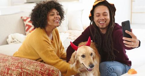 Couple Taking Selfie With Golden Retriever Wearing Red Devil Horns On Cozy Sunlit Living Room Couch