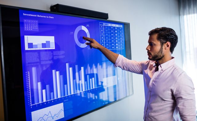 Businessman analyzing data on a touch screen in a modern office. Ideal for use in articles and presentations about business analytics, data-driven decision making, corporate finance, and modern office technology. Suitable for illustrating concepts related to professional work environments, investment analysis, and interactive digital tools.