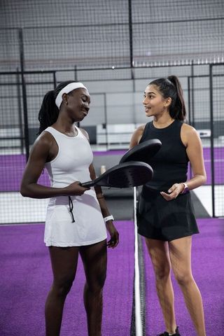 Indian and african american women chatting and holding rackets on purple padel court