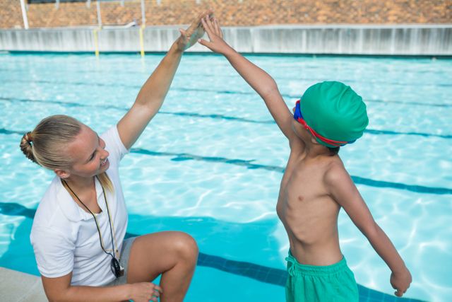 Female trainer giving high five to young boy wearing swim cap and goggles at poolside. Ideal for use in content related to swim training, sports coaching, child development, summer activities, fitness programs, and motivational materials.