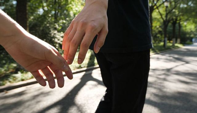 Hands reaching while walking on sunlit tree-lined park path, intimate connection