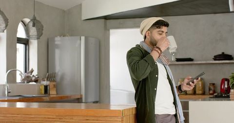 Urban young indian man drinking coffee and checking smartphone at modern kitchen island