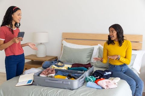 Multicultural friends packing carry-on suitcase on bed preparing weekend getaway together