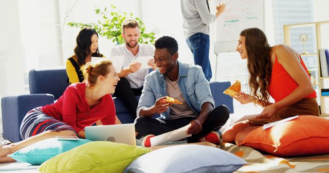 Diverse group of colleagues sitting on pillows in modern office environment, collaborating and laughing while sharing pizza. Perfect for use in materials promoting teamwork, coworking spaces, or modern office culture.