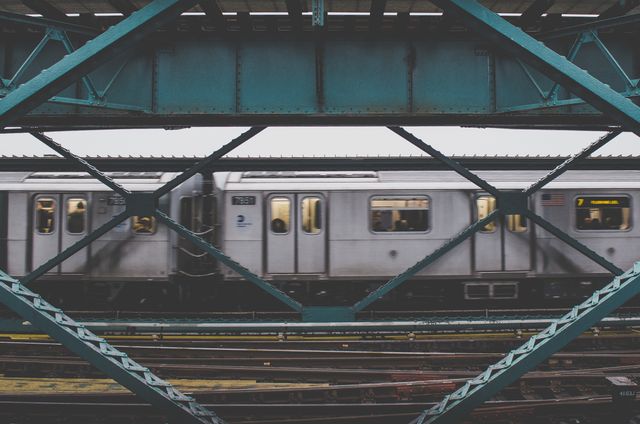 The image shows a subway train moving through a city, framed by the steel beams of a bridge on an overcast day. It captures the essence of urban infrastructure and the daily commute. This image can be used in contexts related to city life, transportation, commuting, and urban development.