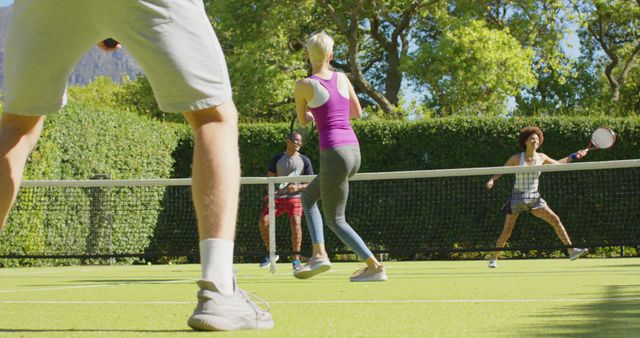 Group of friends playing pickleball on brightly lit court surrounded by greenery. Great for promoting active lifestyles, social recreational activities, or pickleball's growing popularity. Suitable for articles, sports-related blogs, or fitness program advertisements.