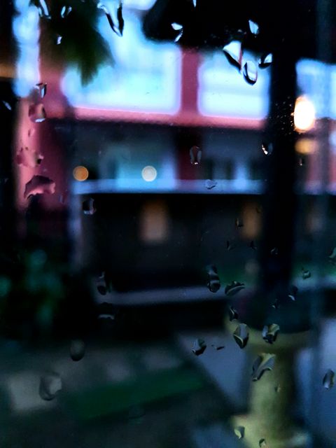 Image showcases raindrops on windowpane with a blurred building out of focus in the background during evening. Use this to evoke themes of tranquility, melancholy or reflections related to rainy weather. Ideal for blog posts, social media, weather-related articles or mood boards.