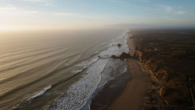Aerial coastline at golden hour showing sea stacks, waves and cliffs along sandy shore