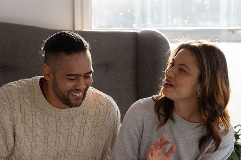 Diverse couple sharing cozy sunlit conversation on grey sofa wearing cable knit sweaters