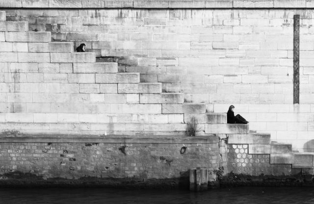 Black and white depiction of two solitary figures sitting on stone steps by a river. Captures an urban setting with stark architectural lines and textures. Can be used for themes of solitude, introspection, urban life, and reflections in photography.
