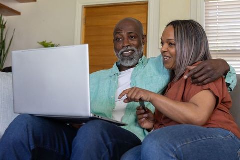 African american couple relaxing on couch using laptop, sharing remote and smiling