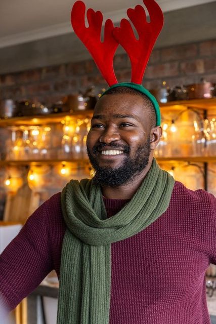 African american man smiling wearing reindeer antlers and green scarf in cozy festive cafe