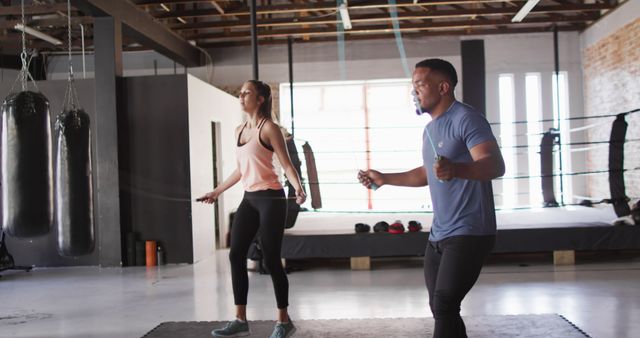 Two individuals are engaged in a fitness workout session, actively skipping rope in an urban gym. The gym features boxing equipment, such as hanging punching bags and a boxing ring. This image is ideal for promoting fitness routines, gym memberships, and active lifestyle content.