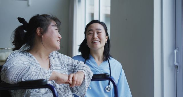 A nurse in light blue scrubs interacting warmly with a patient in a wheelchair by a large window, both sharing a smiling moment. This can be used to represent compassionate healthcare, support services, and patient-nurse relationships in hospitals or clinics. Ideal for websites, brochures, and promotional materials related to medical care, nursing, and senior care.