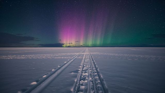 Aurora over frozen plain with parallel snow tracks leading to colorful night sky