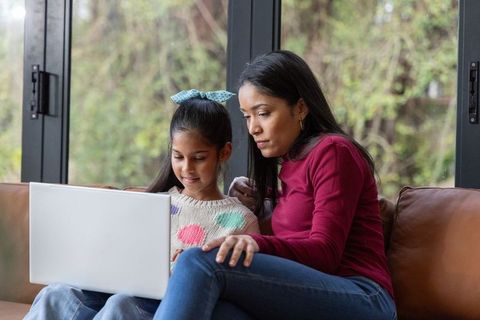 Mother and daughter learning together on laptop while sitting on brown leather sofa by window