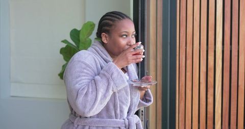 African american woman wearing plush bathrobe sipping tea on sunny balcony morning