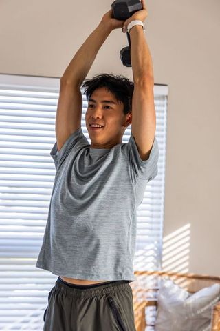 Asian man lifting dumbbell overhead during home workout with natural light and blinds
