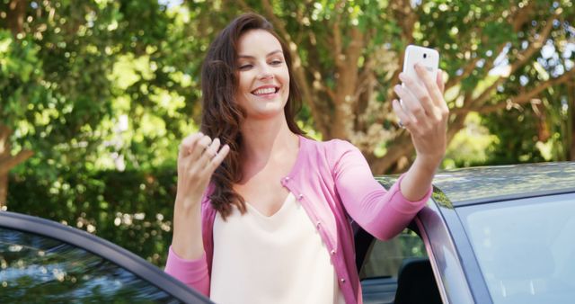 A woman wearing a pink cardigan and white blouse is taking a selfie with her smartphone next to a car in an outdoor setting surrounded by greenery. Ideal for use in automotive, lifestyle, technology, and social media concepts, promoting outdoor activities, or depicting modern connectivity and happiness.