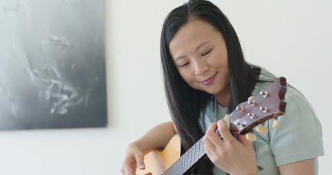 Asian woman playing acoustic guitar, practicing chords and strumming in relaxed interior